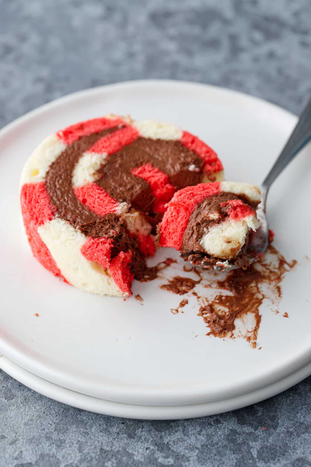 White plate with slice of Peppermint Striped Cake Roll with Chocolate Peppermint Whipped Cream, a messy forkful taken out to show the textures of the cake and filling.