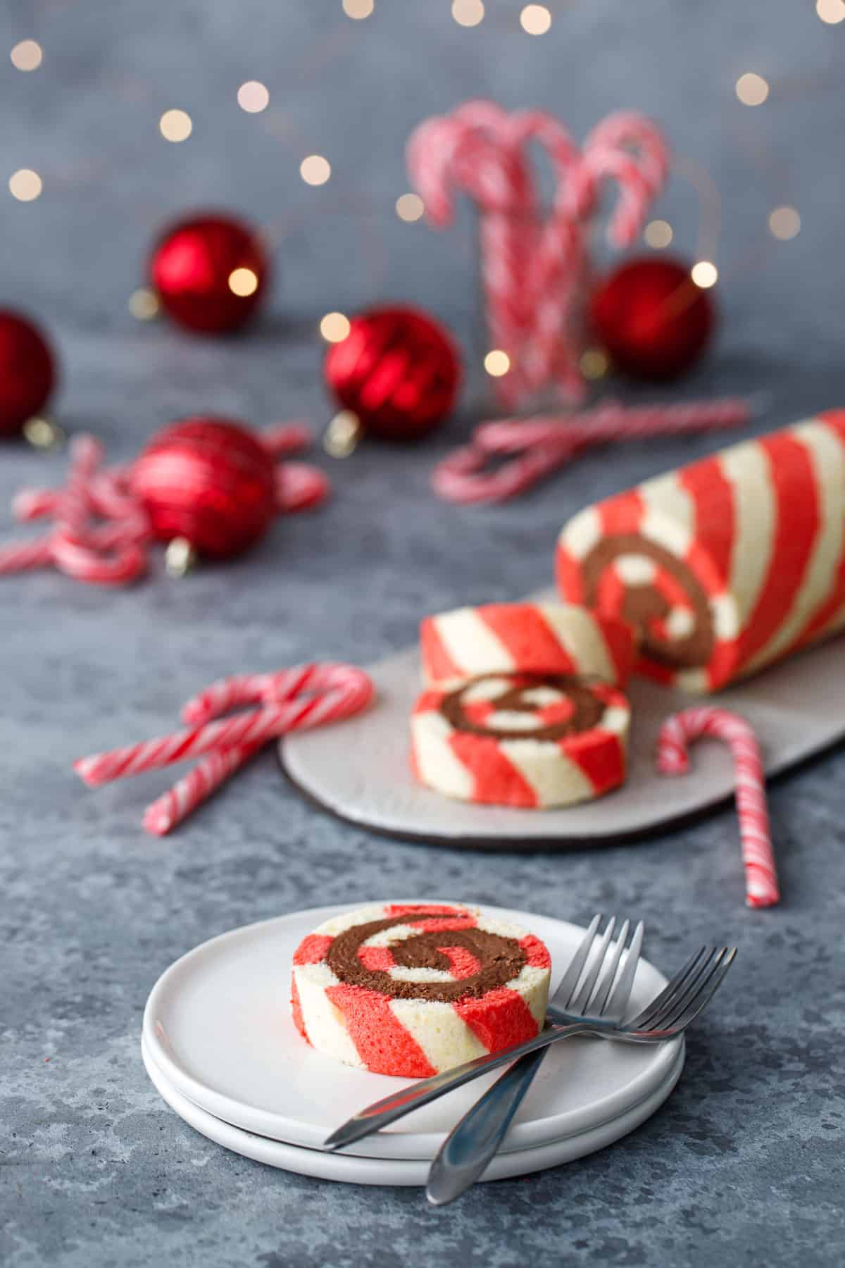 Slice of red and white Peppermint cake roll on a white ceramic plate in the foreground, full cake roll out of focus in the background along with candy canes, ornaments, and Christmas lights.