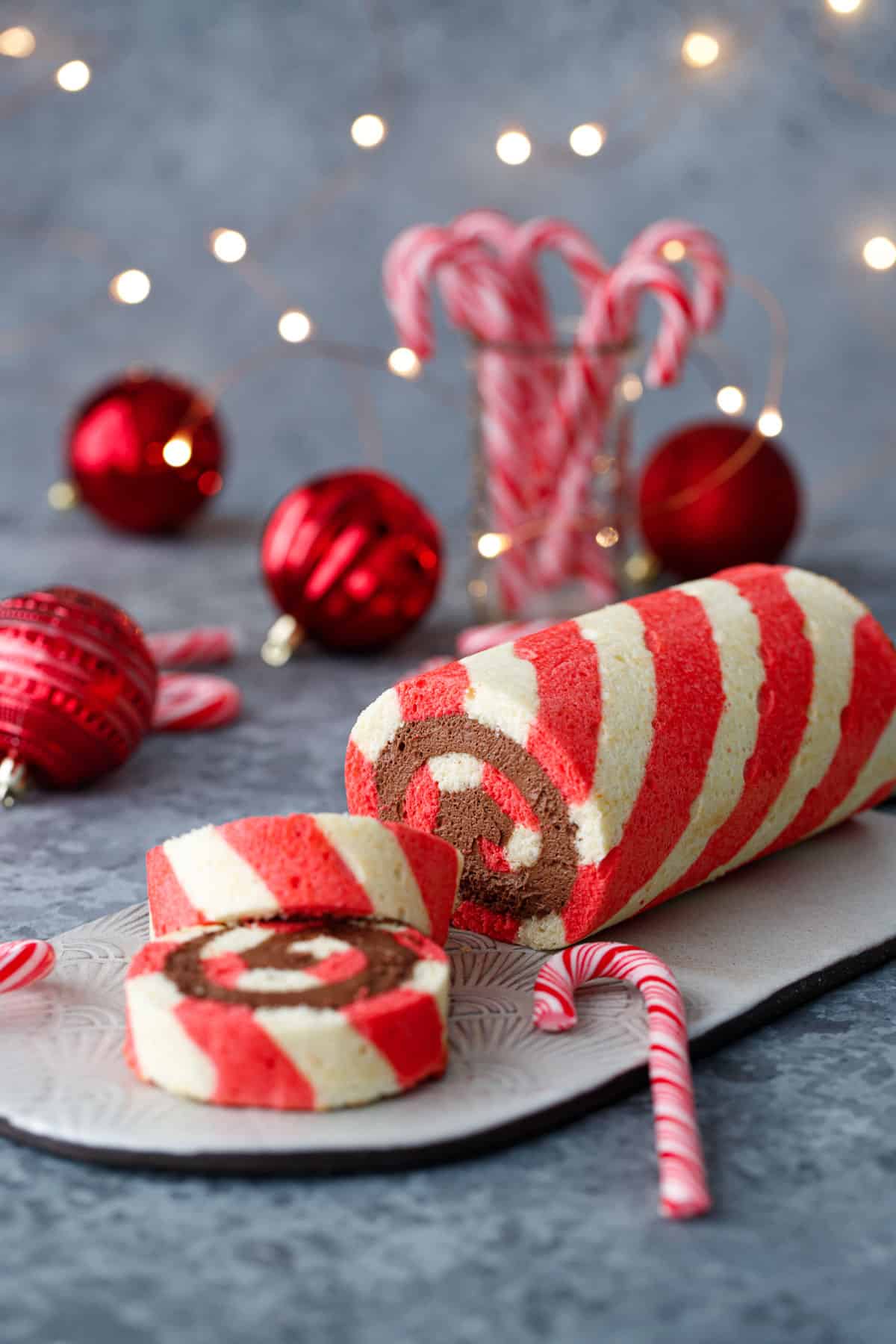 Red and white Peppermint cake roll with two slices cut off and laying down to show a perfect spiral of chocolate peppermint whipped cream filling, with candy canes, ornaments, and Christmas lights out of focus in the background.