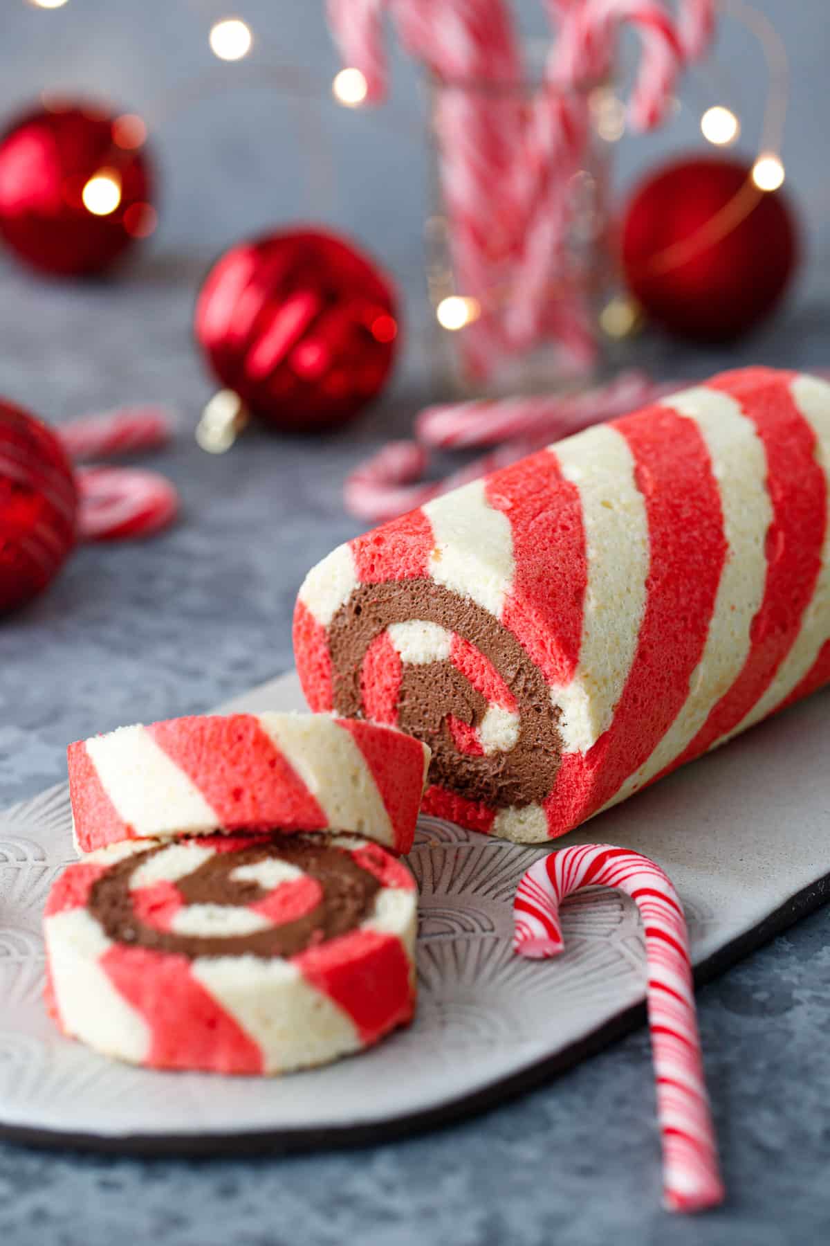Red and white Peppermint cake roll with two slices cut off and laying down to show a perfect spiral of chocolate peppermint whipped cream filling, with candy canes, ornaments, and Christmas lights out of focus in the background.
