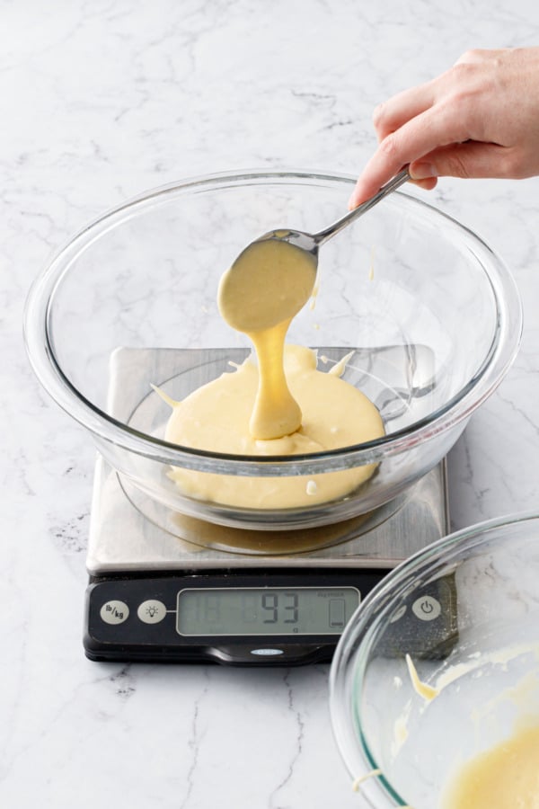 Spooning cake batter into a glass mixing bowl on a kitchen scale.