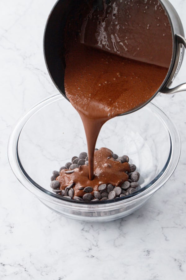Pouring hot cocoa mixture in saucepan over top of glass bowl filled with chocolate callets.