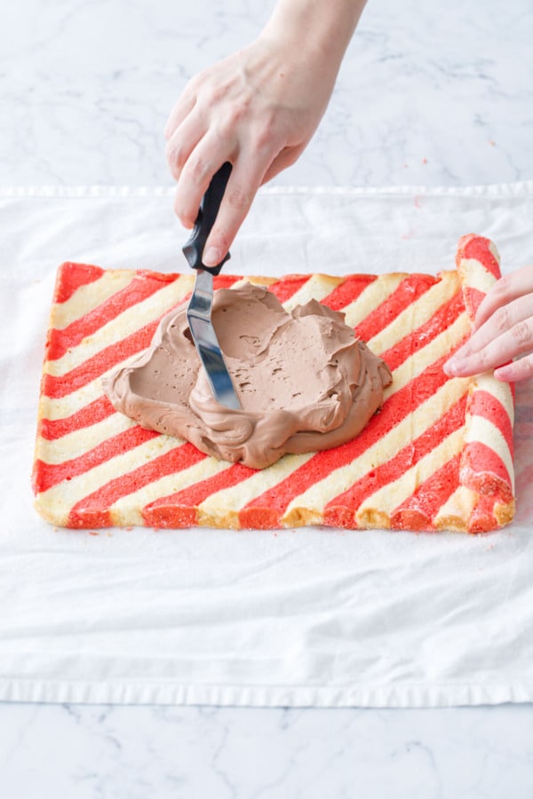 Spreading chocolate whipped cream filling onto the unrolled striped cake with an offset spatula.