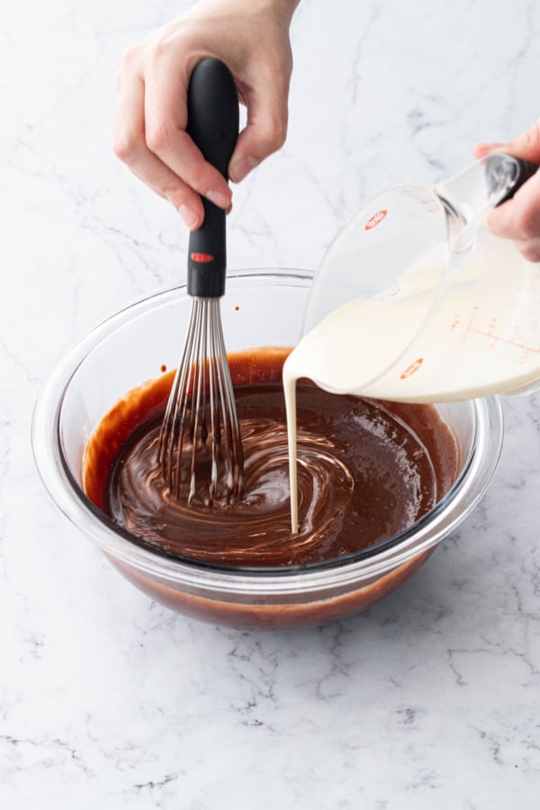 Whisking chocolate mixture in bowl while pouring in heavy cream from a measuring cup.