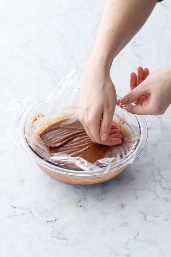 Pressing a layer of plastic wrap down onto the surface of the melted chocolate mixture to prevent a skin from forming.