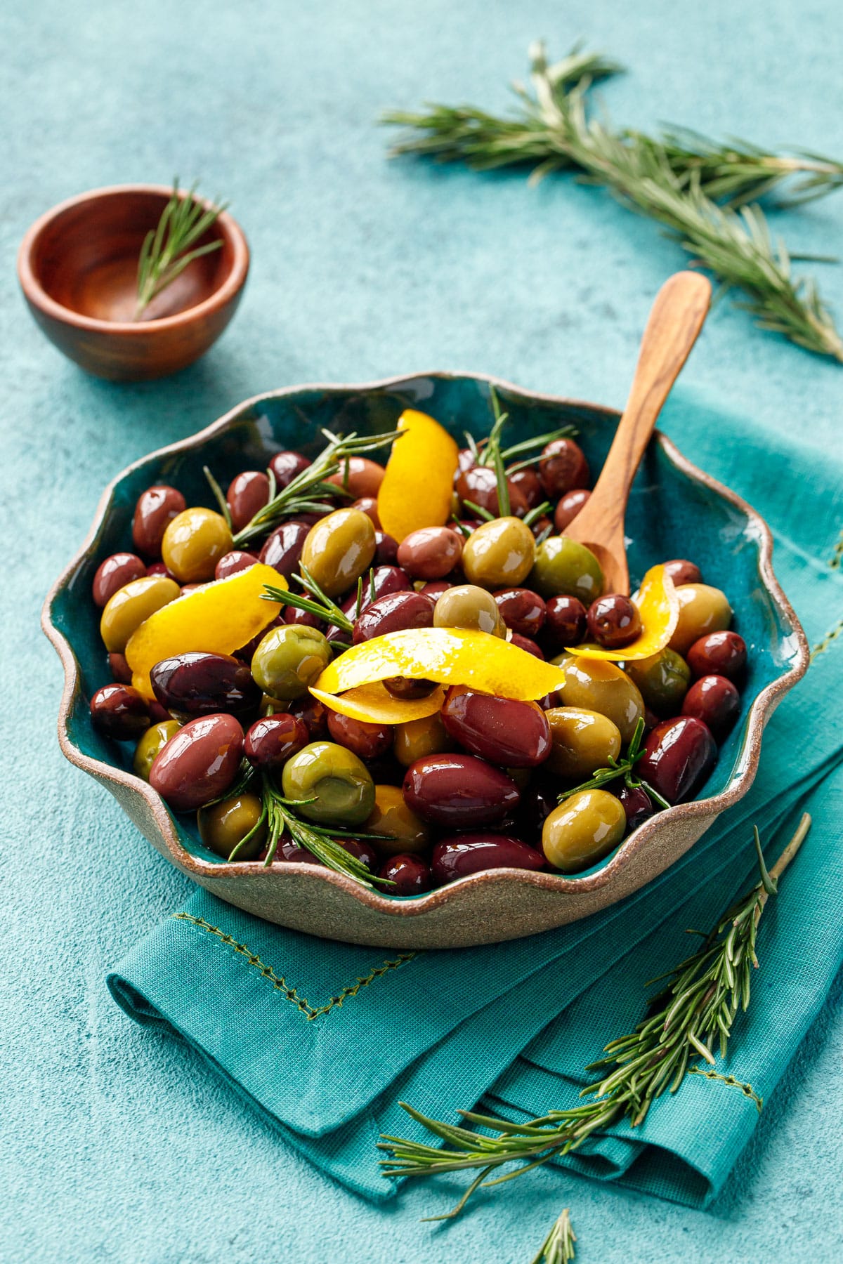 Turquise ceramic dish with warm mixed color olives, shiny with pieces of lemon peel and fresh rosemary, on a turquoise background.