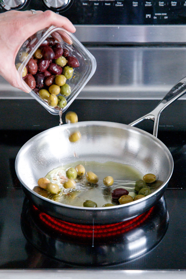 Pouring multi-colored olives from a container into a pre-heated skillet on the stove.