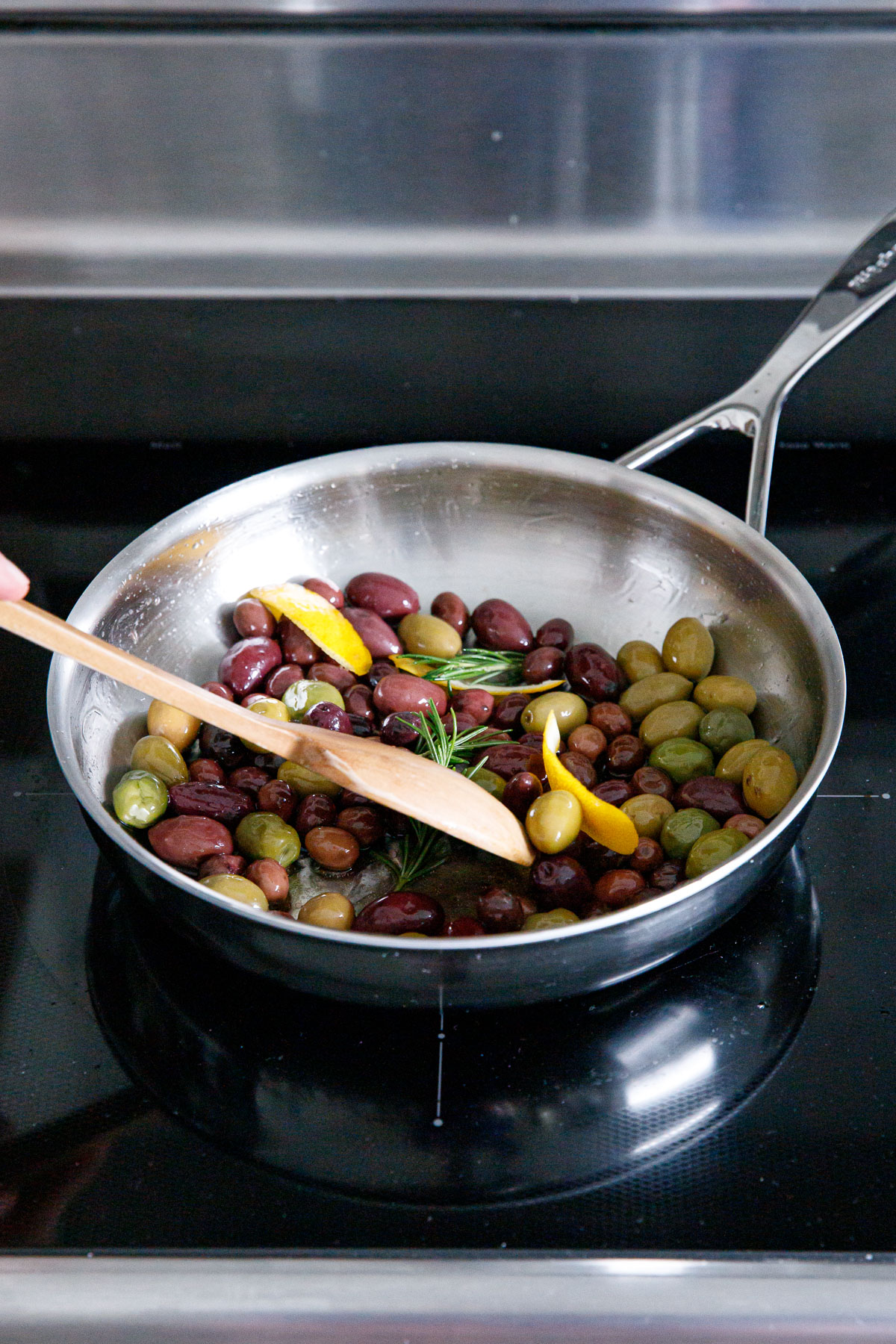 Wooden spoon stirring multi-colored olives in a skillet with strips of lemon peel and rosemary.