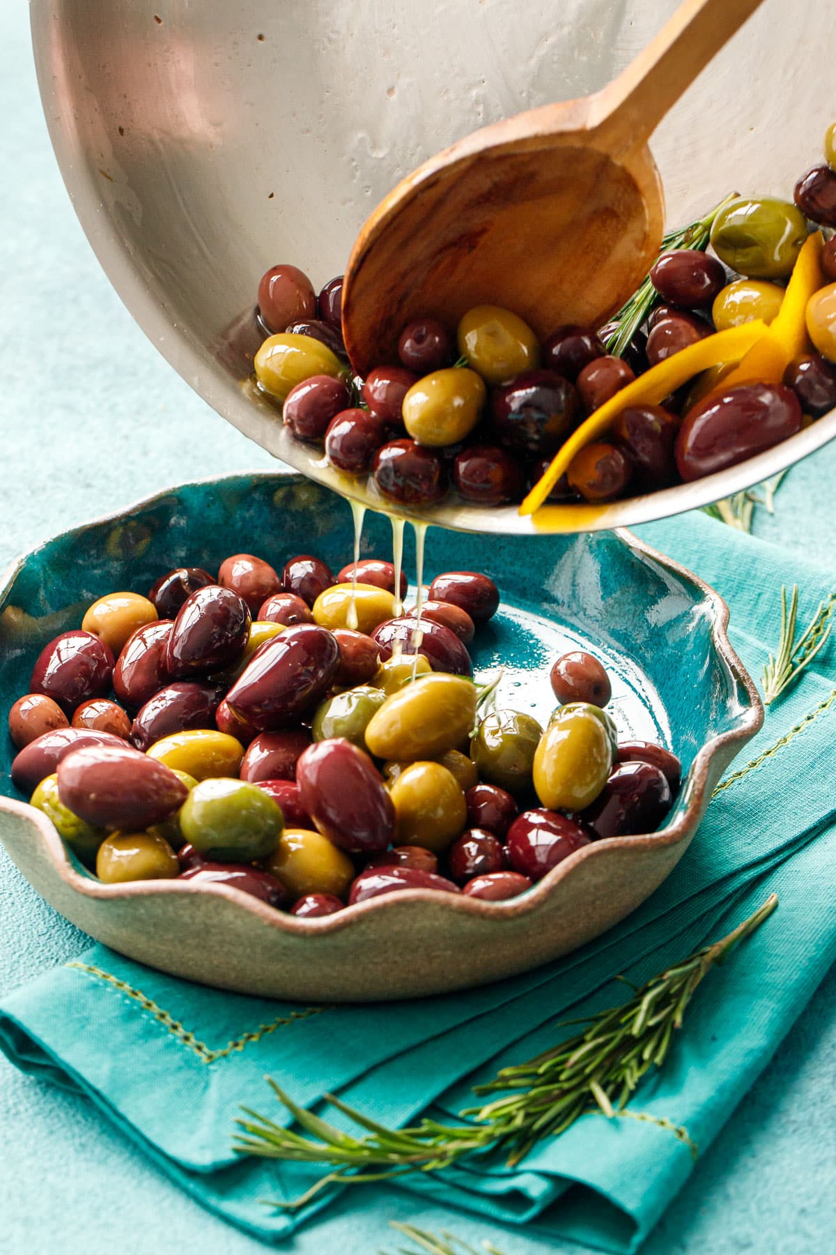 Pouring warm sautéed olives from stainless skillet into a ruffled-edge turquoise ceramic bowl with visible drips of oil.
