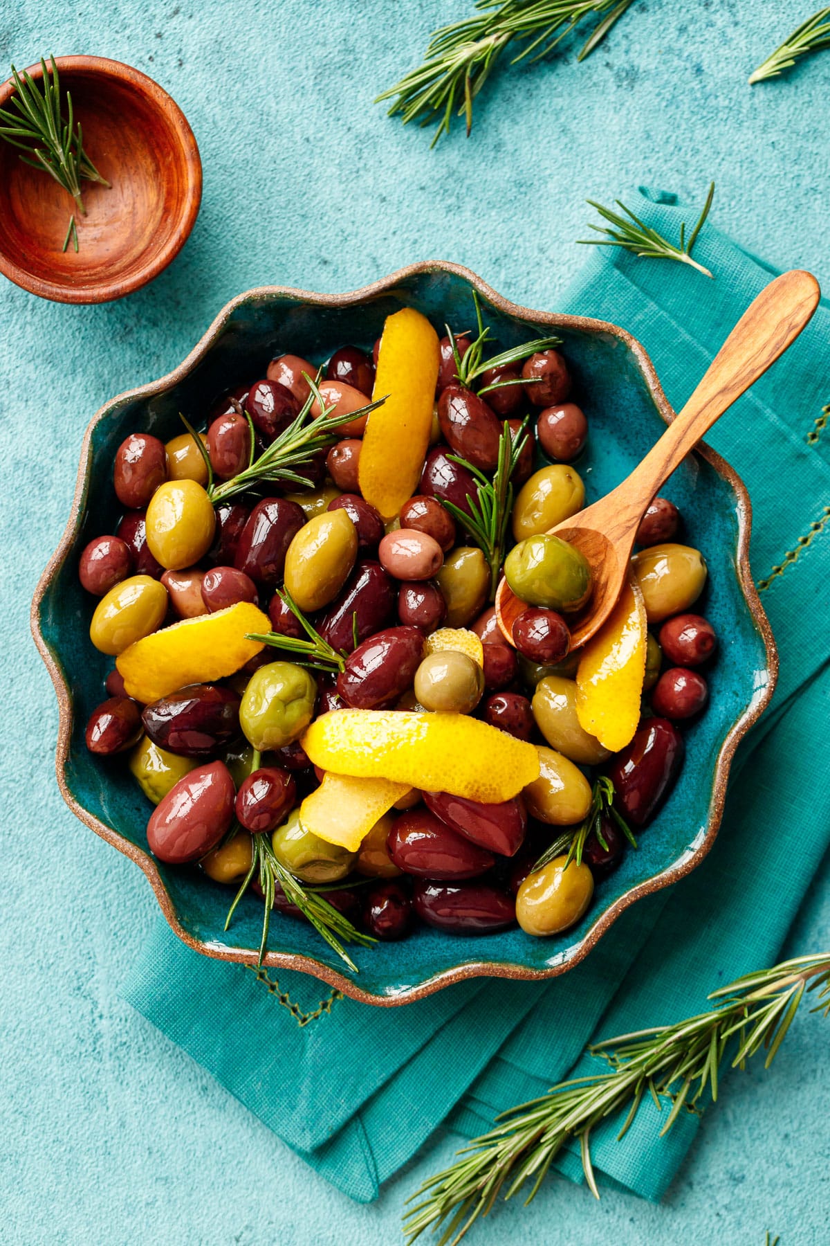 Overhead, ruffled edge bowl filled with shiny multi-colored olives sauteed with strips of lemon peel and fresh rosemary, on a turquoise background.