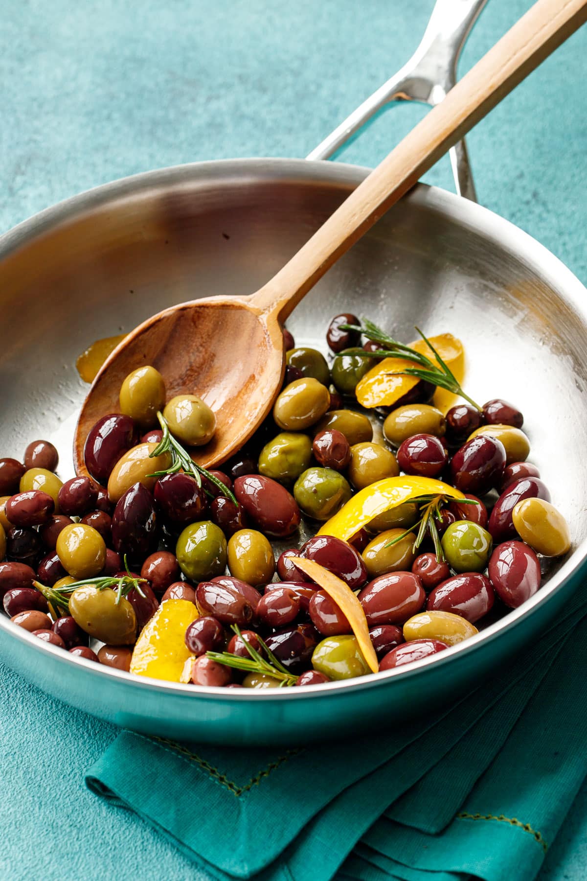 Stainless steel skillet with warm sauteed olives, shiny coated in oil with pieces of lemon peel and fresh rosemary and a wooden spoon.