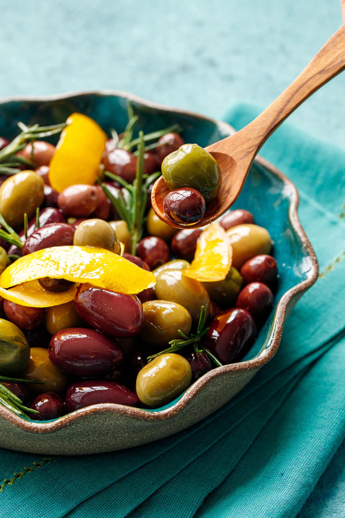 Closeup, small wooden spoon lifting one purple and one green olive above a bowl filled with shiny sauteed olives with pieces of lemon peel and rosemary.