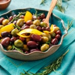 Mixed purple and green olives sauteed and shiny with strips of lemon peel and fresh rosemary, with a small wooden spoon in a turquoise ceramic bowl.