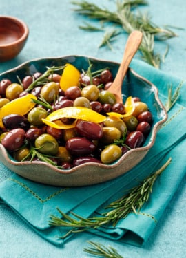 Mixed purple and green olives sauteed and shiny with strips of lemon peel and fresh rosemary, with a small wooden spoon in a turquoise ceramic bowl.