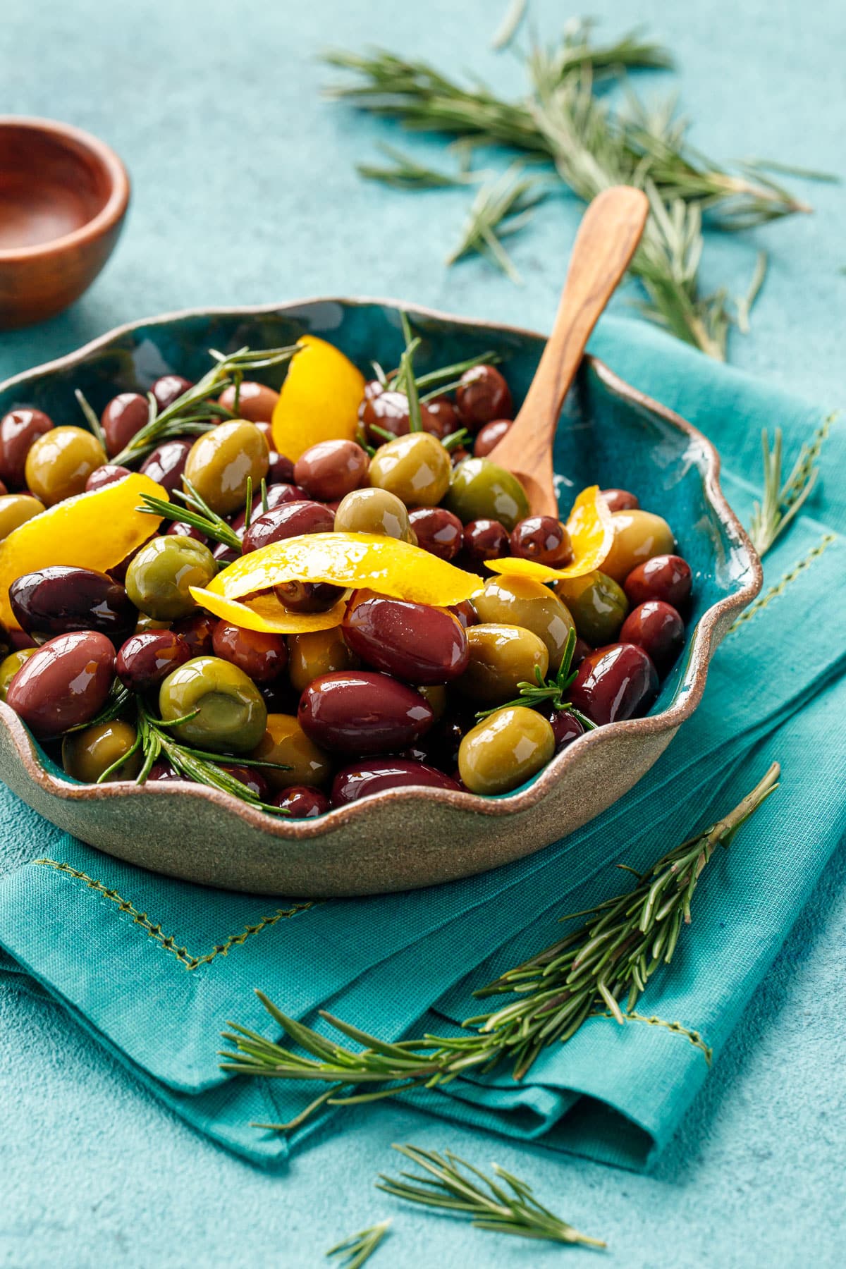 Mixed purple and green olives sauteed and shiny with strips of lemon peel and fresh rosemary, with a small wooden spoon in a turquoise ceramic bowl.