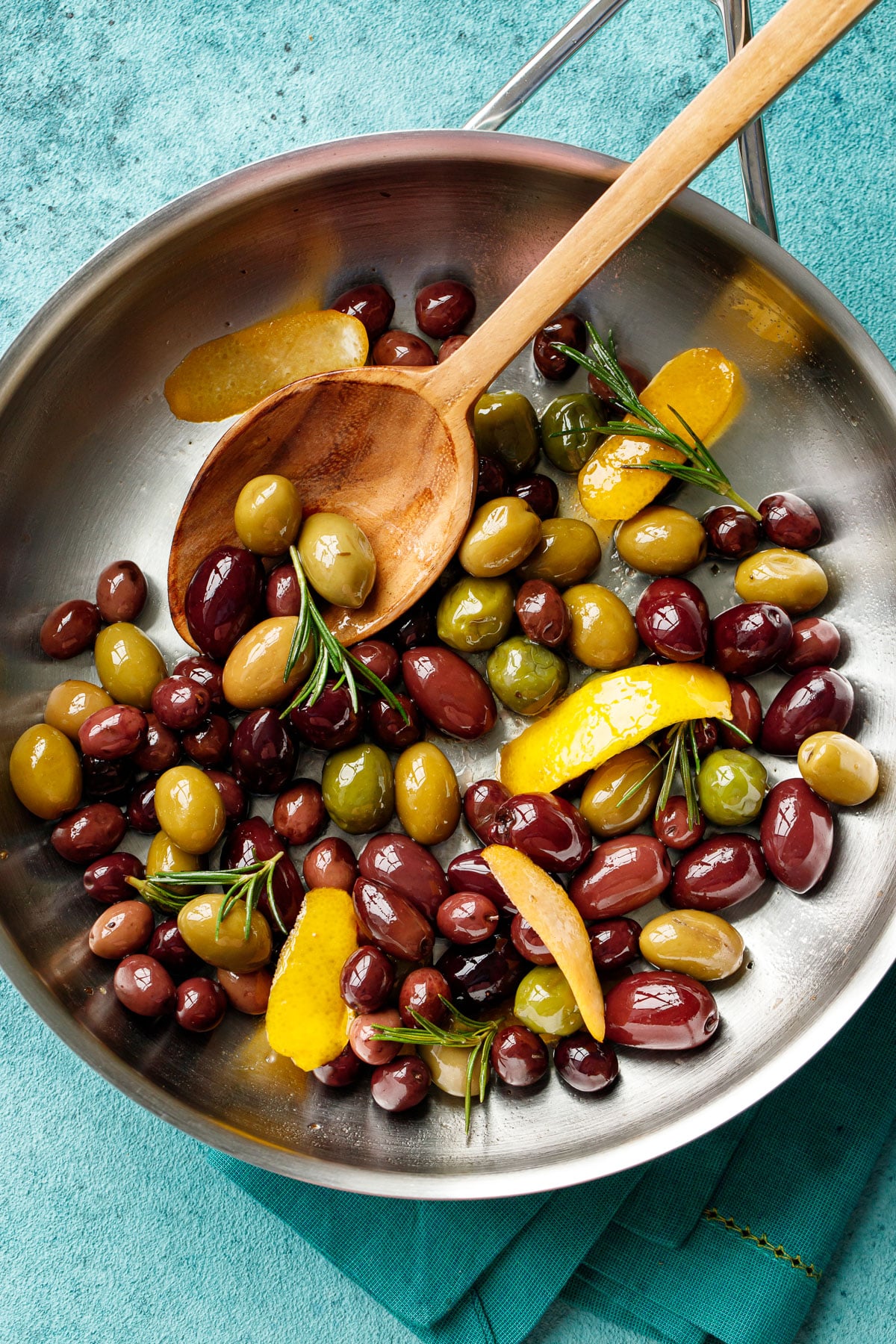 Overhead, stainless steel skillet with shiny warm sautéed olives with visible strips of lemon peel and sprigs of rosemary and a wooden spoon on a turquoise background.