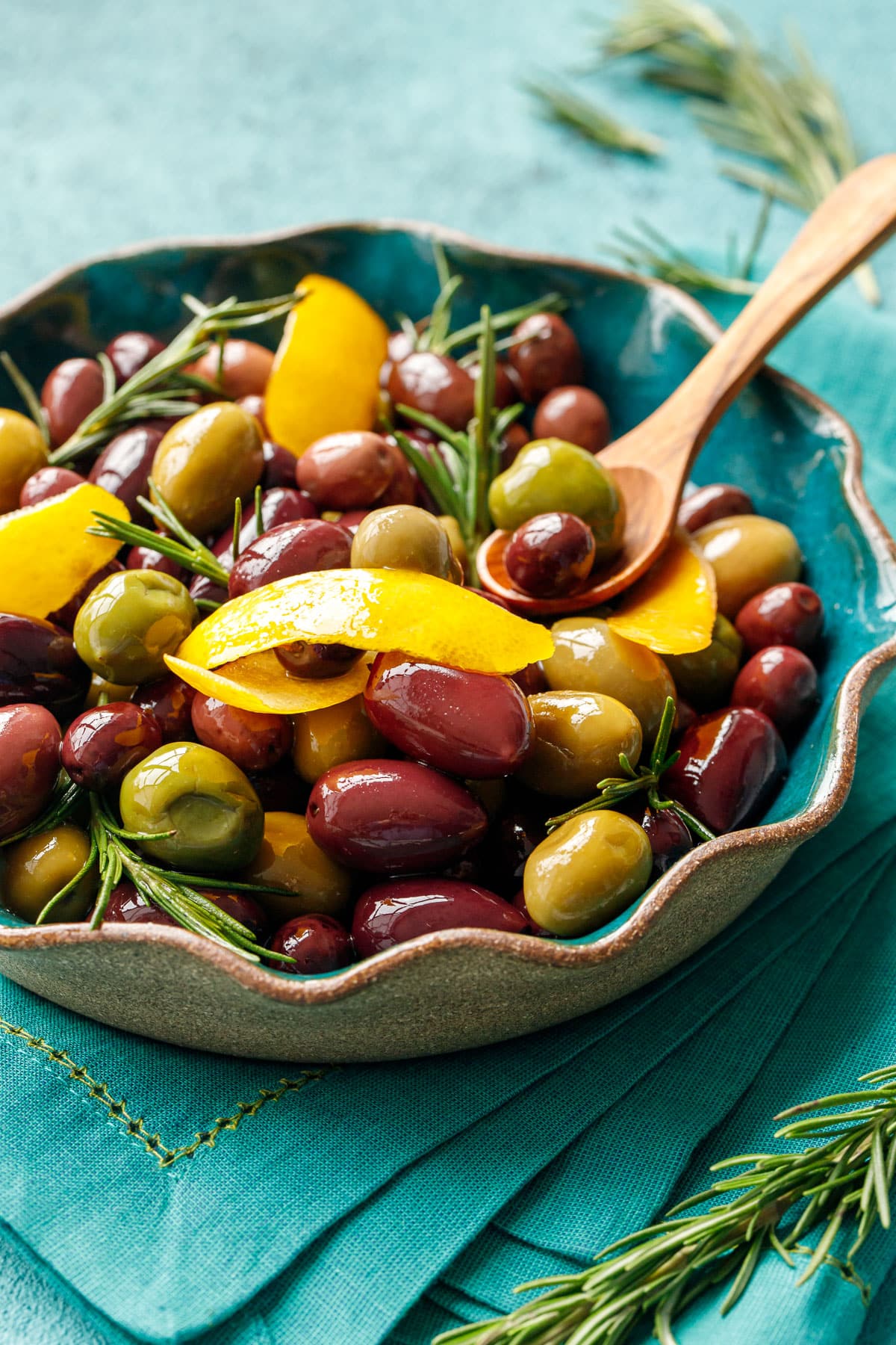 Ruffled turquoise dish with Warm Sautéed Olives with Lemon & Rosemary and a small wooden spoon, fresh rosemary sprigs scattered around.