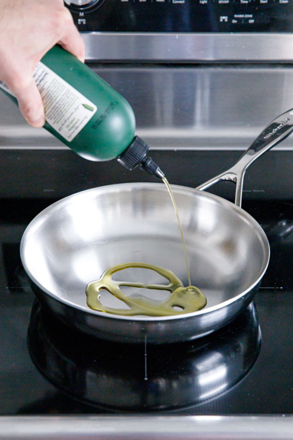 Squirting olive oil from a squeeze bottle into a stainless steel skillet on the stovetop.