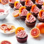 Backlit brightly colored Chocolate Olive Oil & Blood Orange Cupcakes with one cupcake in the foreground and more cupcakes on a wire rack in the background, with fresh blood oranges, candied oranges, and frosting.