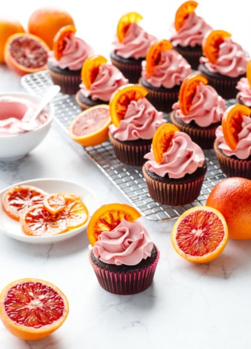 Backlit brightly colored Chocolate Olive Oil & Blood Orange Cupcakes with one cupcake in the foreground and more cupcakes on a wire rack in the background, with fresh blood oranges, candied oranges, and frosting.