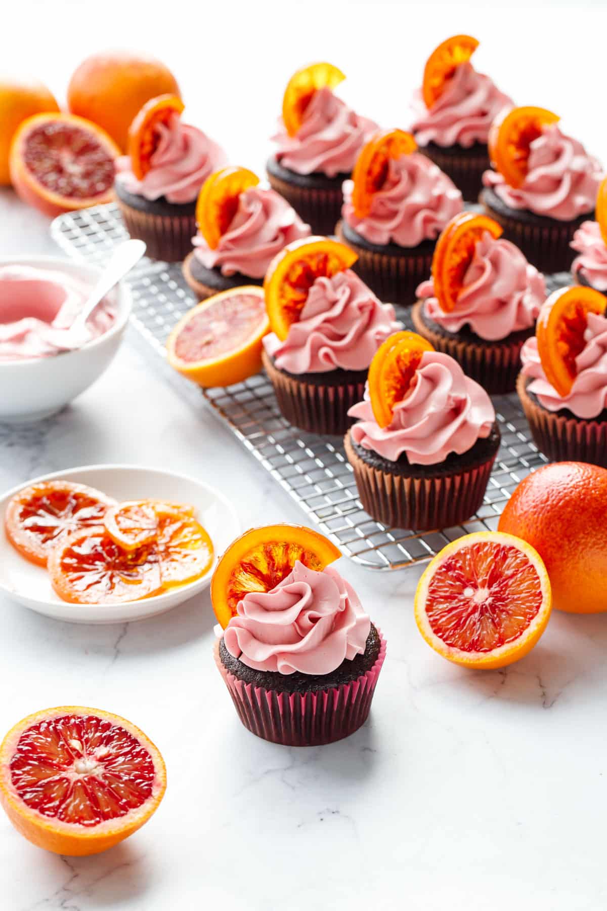 Backlit brightly colored Chocolate Olive Oil & Blood Orange Cupcakes with one cupcake in the foreground and more cupcakes on a wire rack in the background, with fresh blood oranges, candied oranges, and frosting.