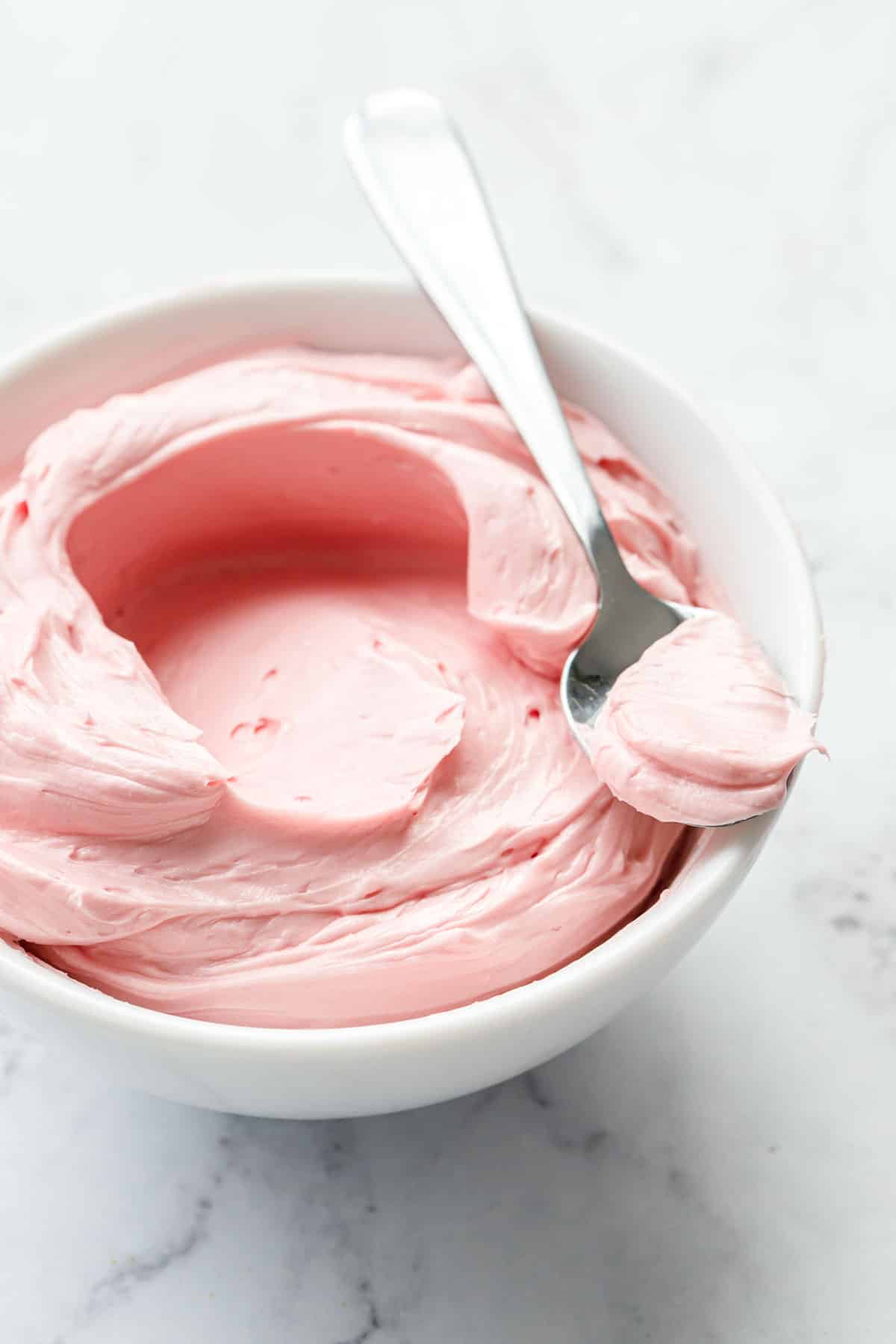 White bowl with swirl of light pink blood orange buttercream frosting, with a small spoon resting on top.