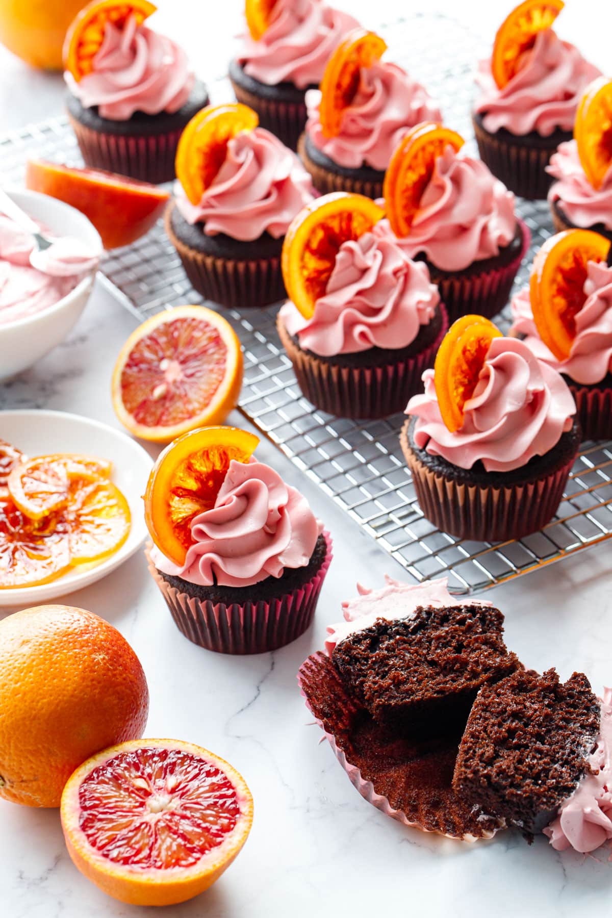 Scattered Chocolate Olive Oil & Blood Orange Cupcakes on a marble background, one cupcake cut in half to show the interior texture, with oranges (fresh and candied), and a bowl of frosting in the background.