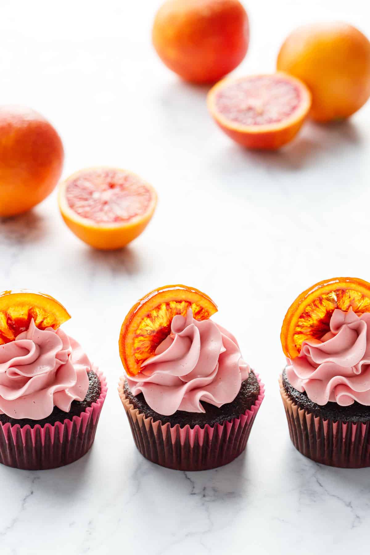 Row of three Chocolate Olive Oil & Blood Orange Cupcakes, backlit and topped with candied orange slices, with fresh blood oranges in the background.