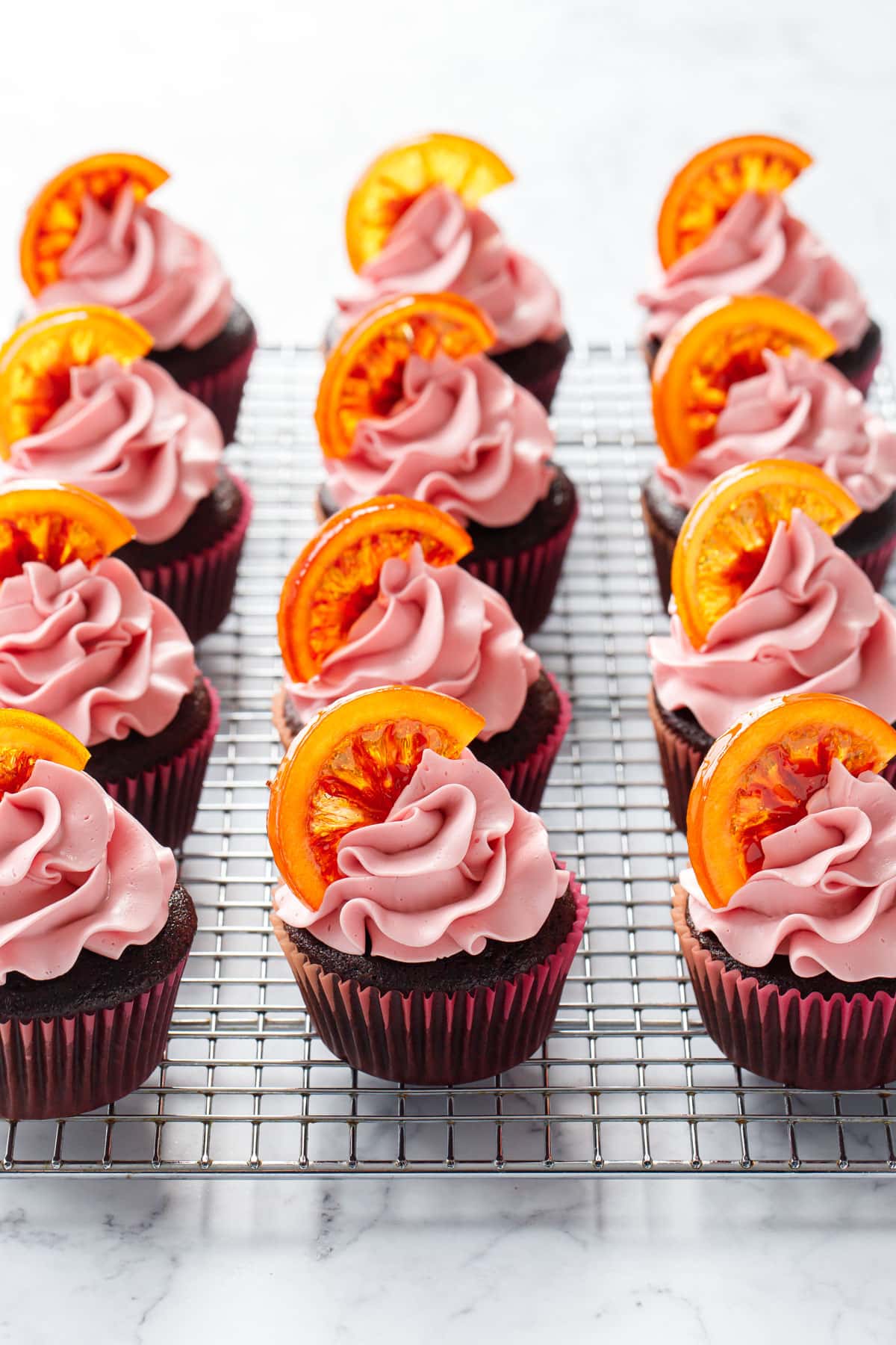 Wire rack with even rows of Chocolate Olive Oil & Blood Orange Cupcakes topped with light pink blood orange buttercream and half slices of candied blood orange.