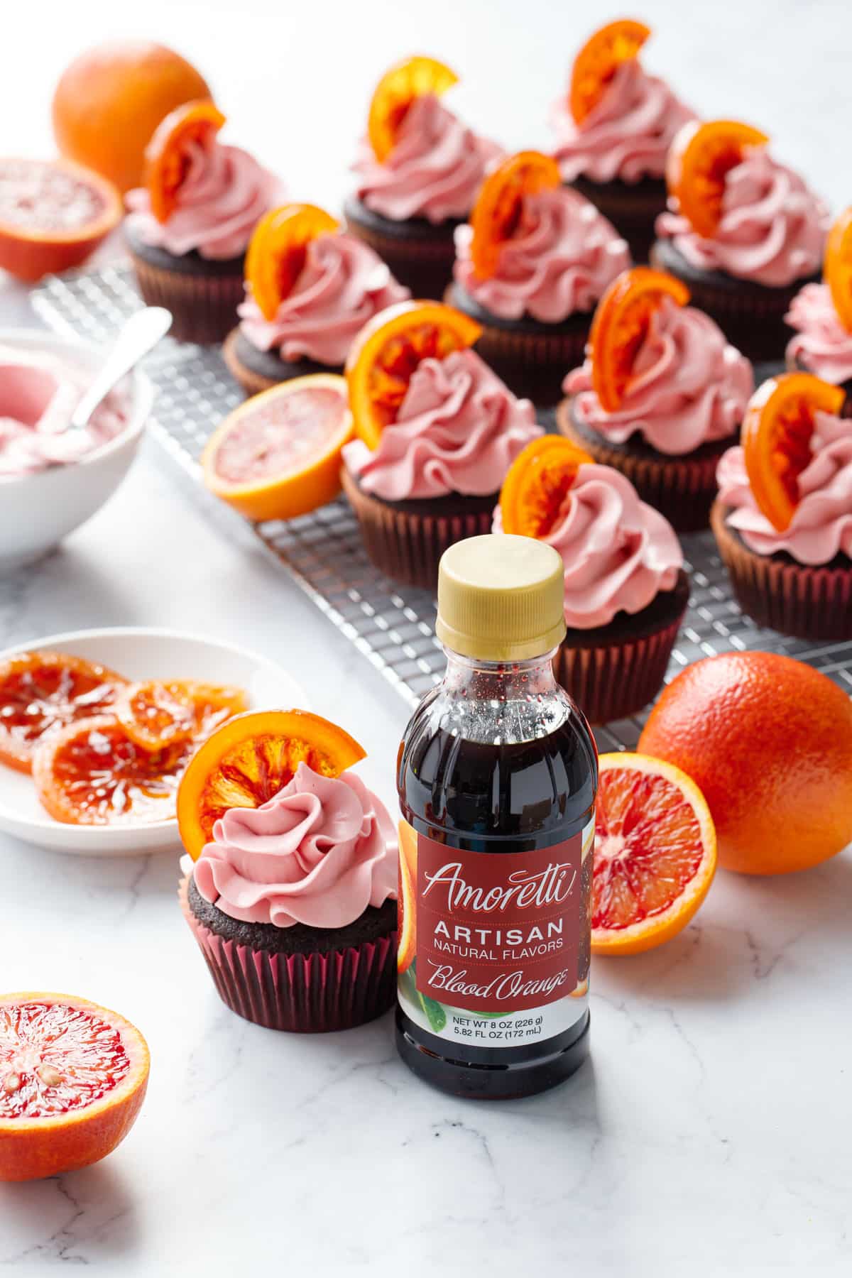 Bottle of Amoretti Blood Orange Artisan Flavoring with Chocolate Olive Oil & Blood Orange Cupcakes on the side and more on a wire rack behind the bottle along with some fresh blood oranges.