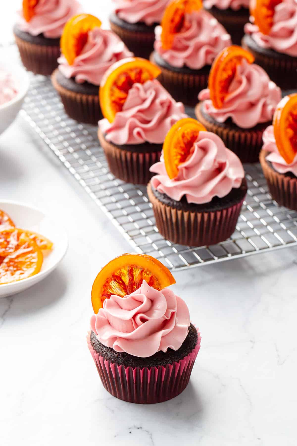 Chocolate Olive Oil & Blood Orange Cupcakes on a marble background, with a cooling rack topped with neat rows of more cupcakes in the background.