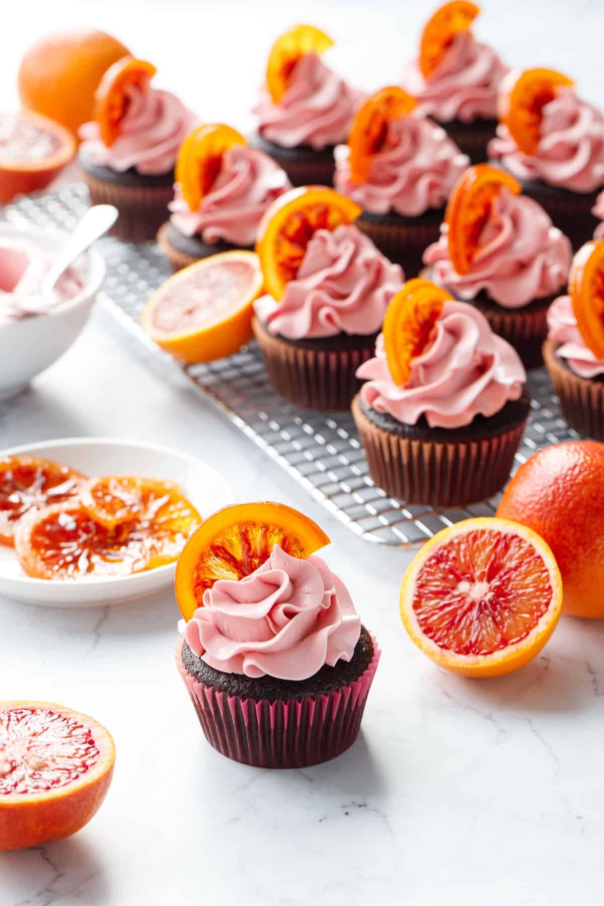 Brightly colored and backlit Chocolate Olive Oil & Blood Orange Cupcakes on a marble background, with fresh blood oranges (whole and halved), plus a plate of candied oranges and bowl of light pink frosting in the background.
