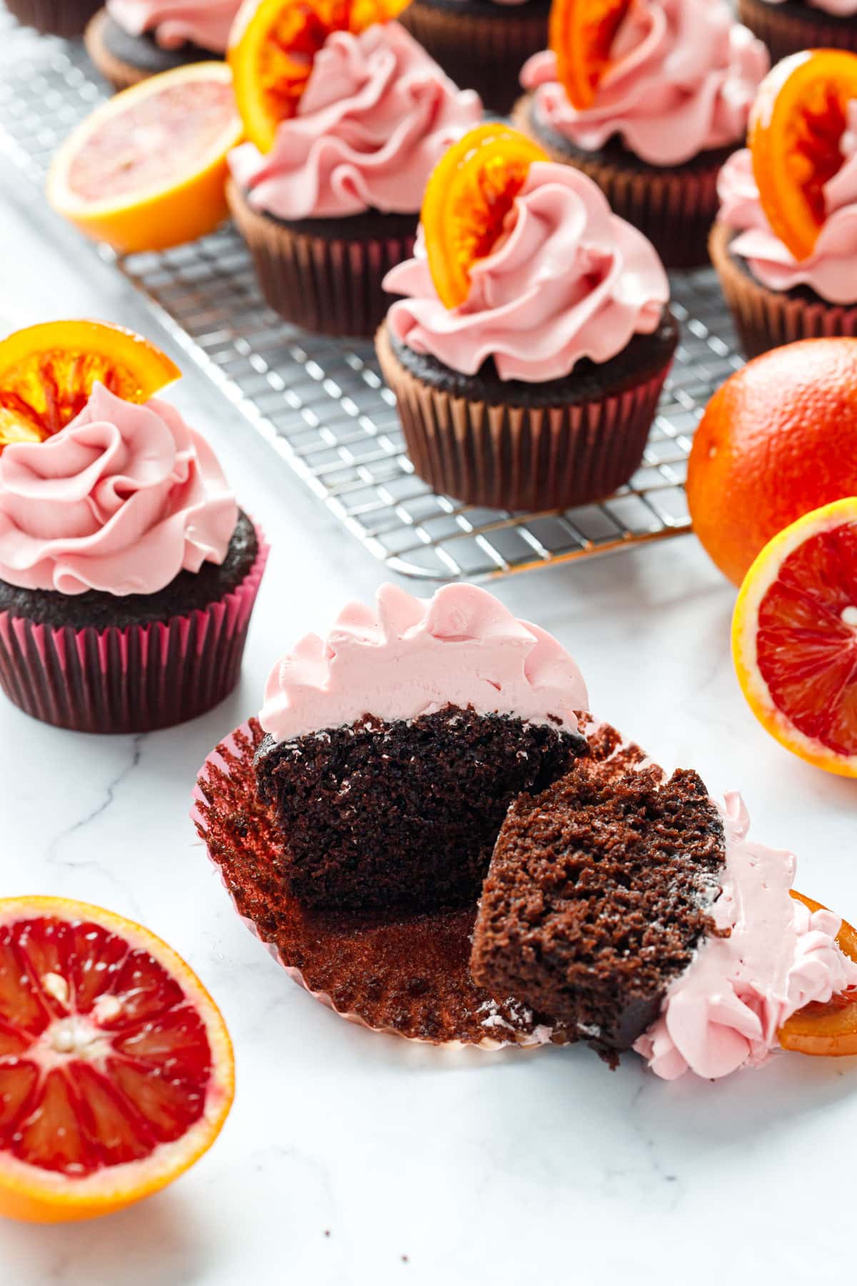 One Chocolate Olive Oil & Blood Orange Cupcake cut in half to show the interior texture of the chocolate cupcake, with a few more cupcakes and some cut blood oranges surrounding it.