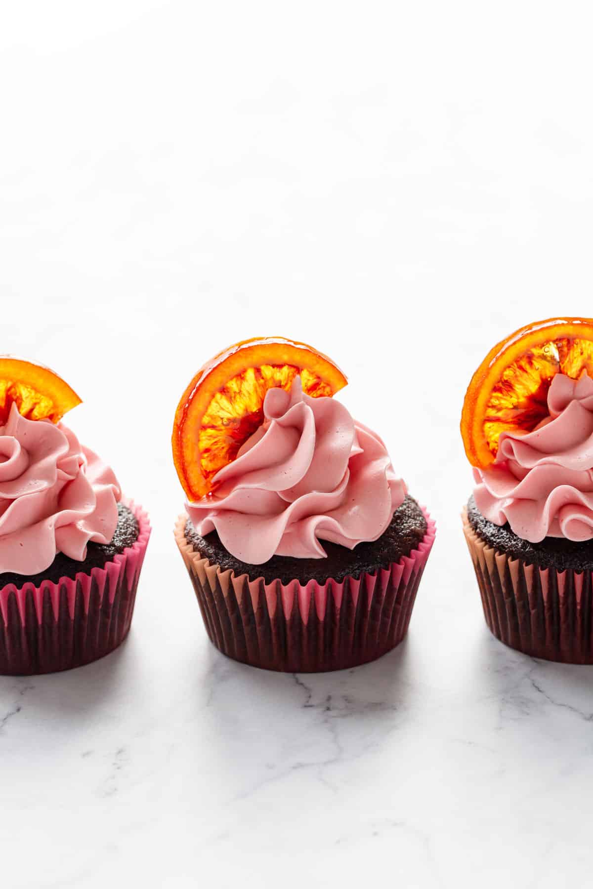 Row of three Chocolate Olive Oil & Blood Orange Cupcakes, backlit on a white marble background, the light shining through the transparent orange slice decorations.