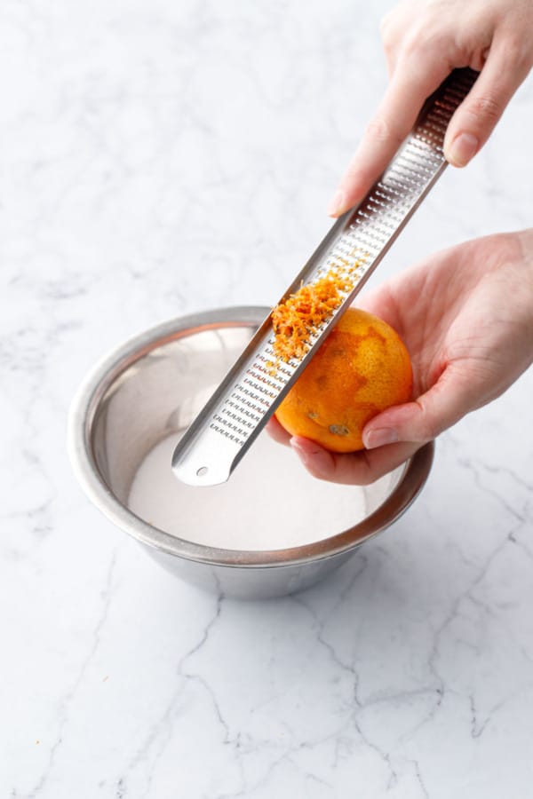 Microplane grating the zest of a blood orange over a bowl of granulated sugar.