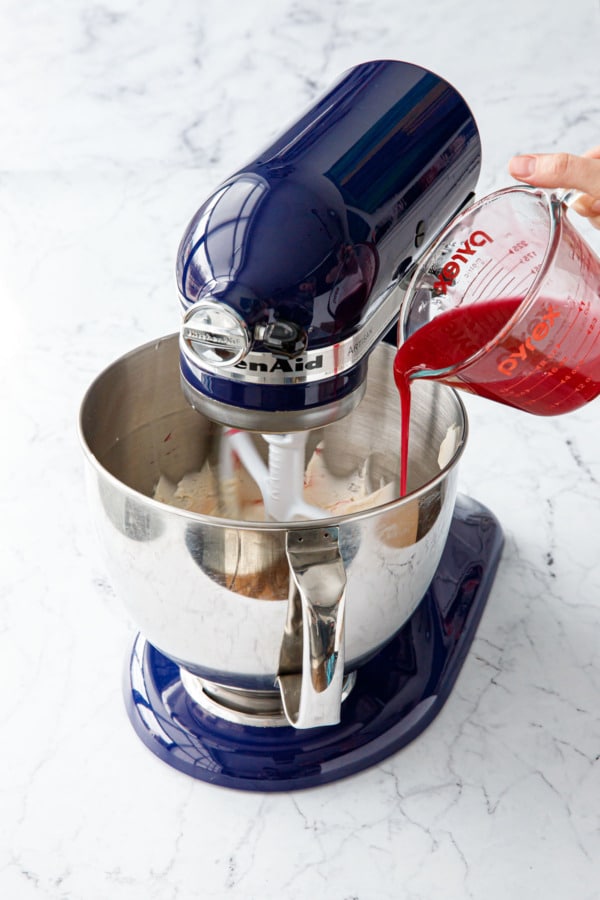 Pouring sugar syrup into whipped butter in a stand mixer on low speed.