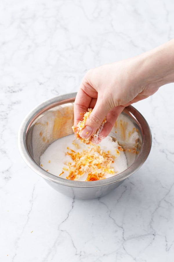 Fingers rubbing orange zest with sugar in a small metal mixing bowl.