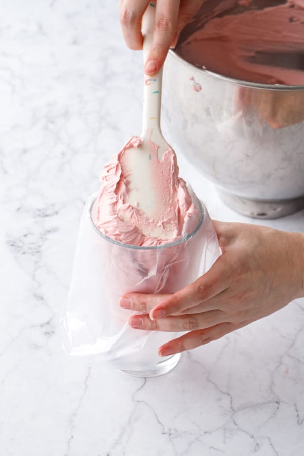 Spatula smushing finished light pink frosting into a piping bag propped up in a drinking glass.