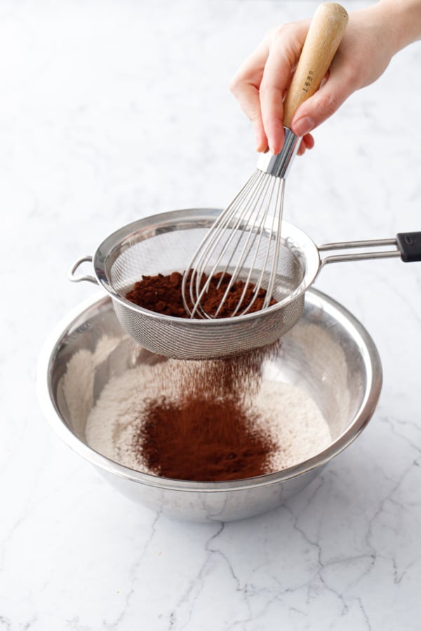 Sifting cocoa powder through a fine mesh sieve into a mixing bowl with flour and dry ingredients.