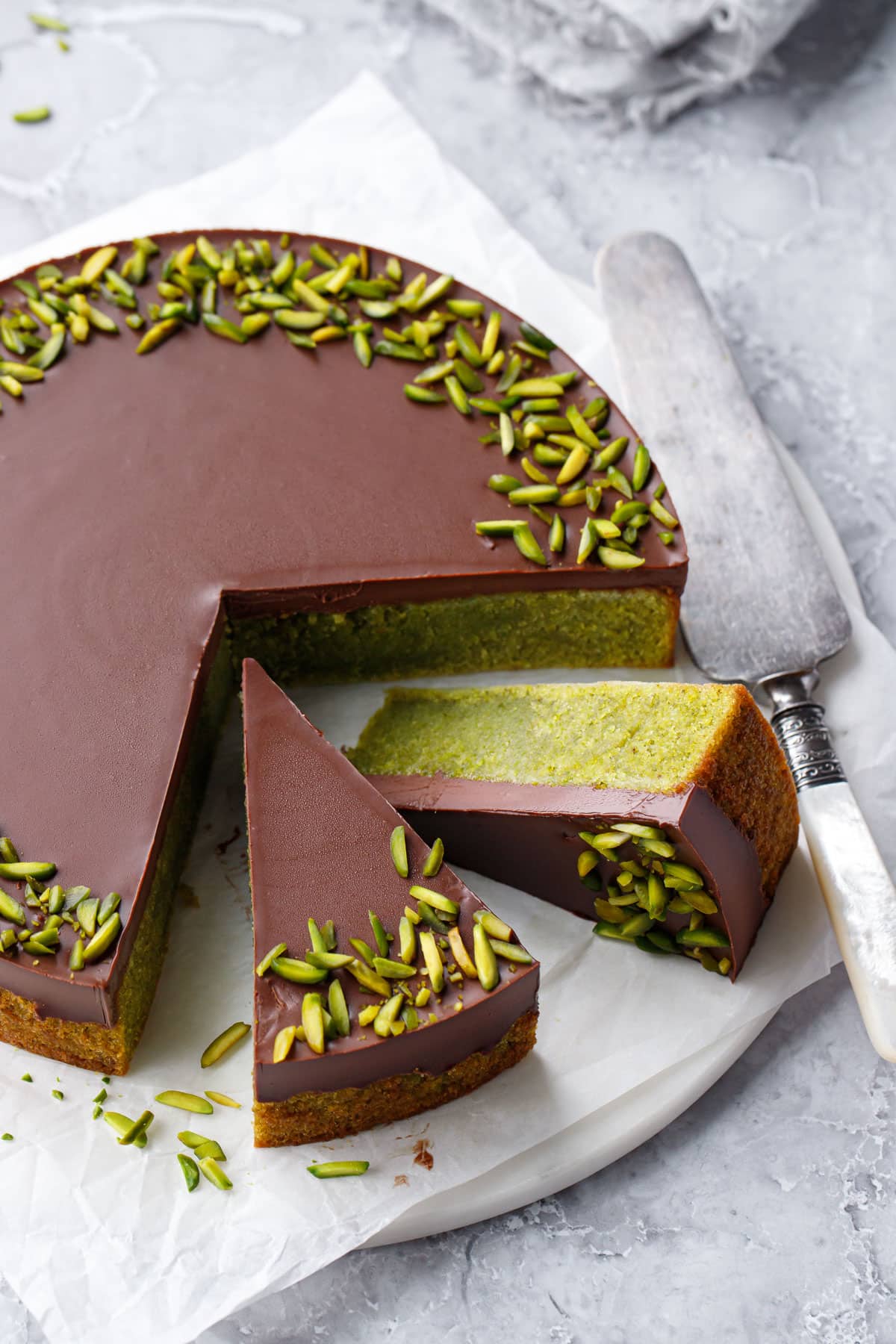 Flourless Pistachio Cake topped with Chocolate Ganache and decorated with slivered pistachios on a flat plate with vintage cake server.