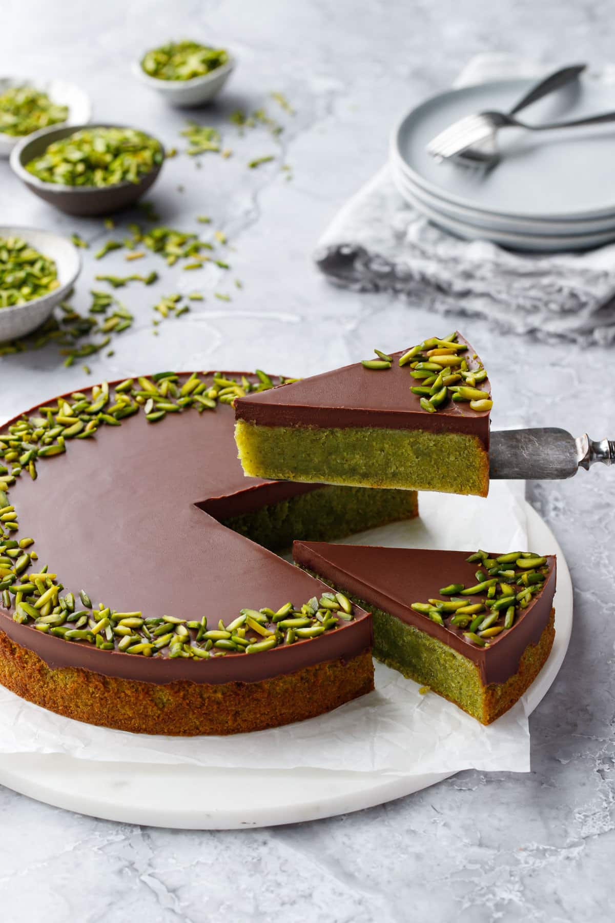 Cake server lifting a cut slice of Flourless Pistachio Cake with a bright green interior and a layer of chocolate ganache on top.