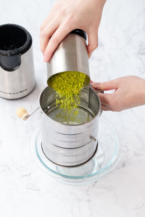 Pouring ground pistachios out of grinder and into a metal sifter set over a glass bowl.