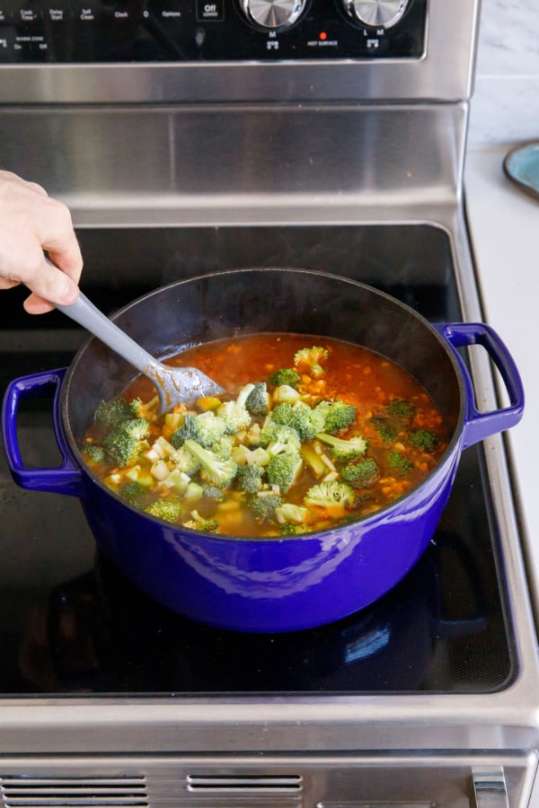 Stirring Brothy Broccoli & Potato Soup in a blue Dutch oven on a glass stovetop.