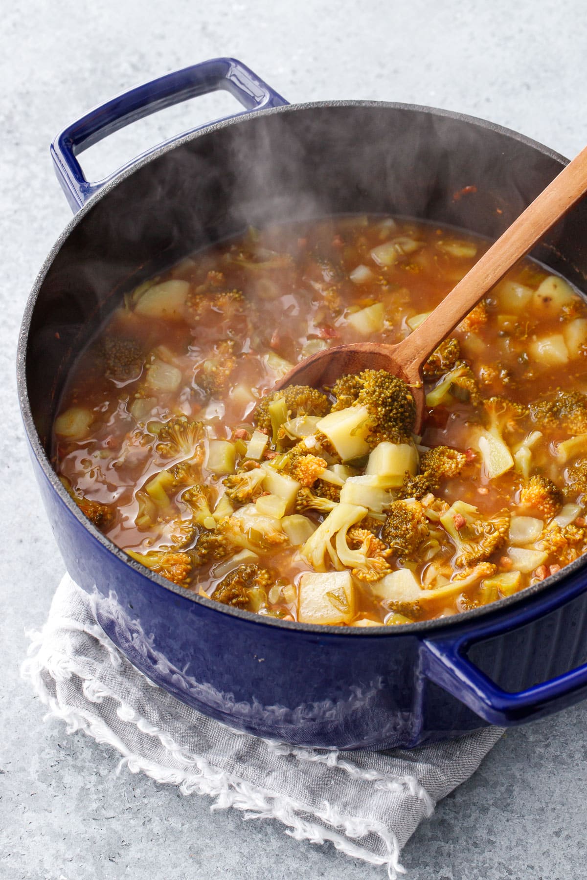 Large blue dutch oven filled with Brothy Broccoli & Potato Soup with a wooden spoon, gray napkin on a gray background.