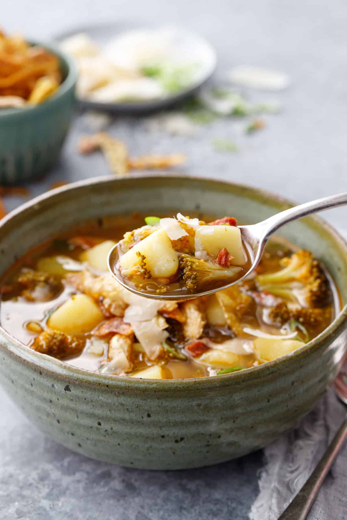 Spoonful of Brothy Broccoli & Potato Soup lifting out of a green ceramic soup bowl, small bowls of toppings in the background.