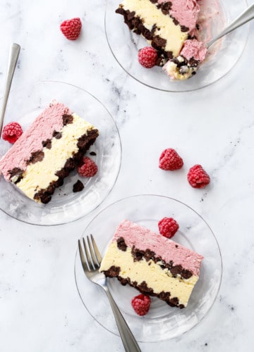 Overhead, marble background and three glass plates with cross-section slices of Raspberry & Passionfruit Semifreddo with Chocolate Crumbs, and a few frozen raspberries scattered around.