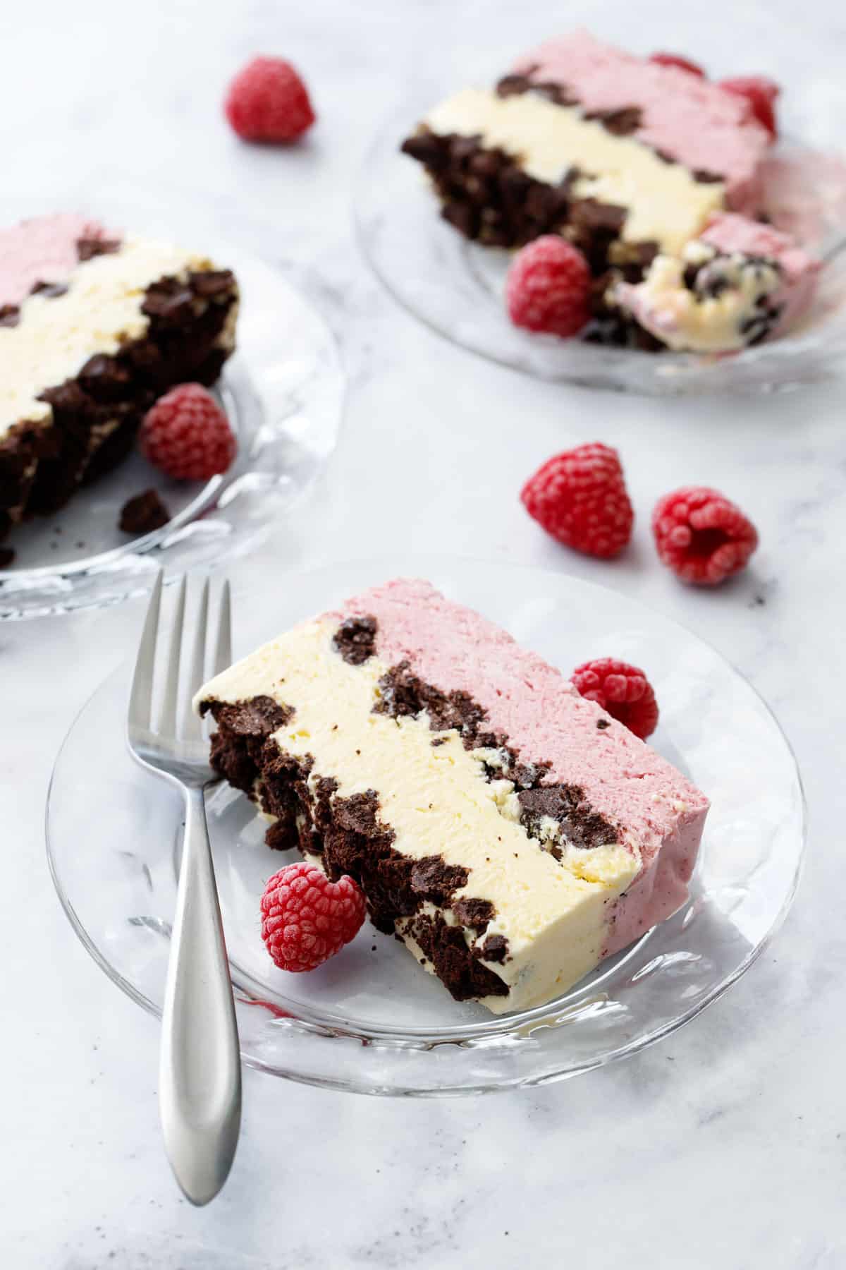 Slices of Raspberry & Passionfruit Semifreddo with Chocolate Crumbs on glass plates with forks and frozen raspberries on a marble background.