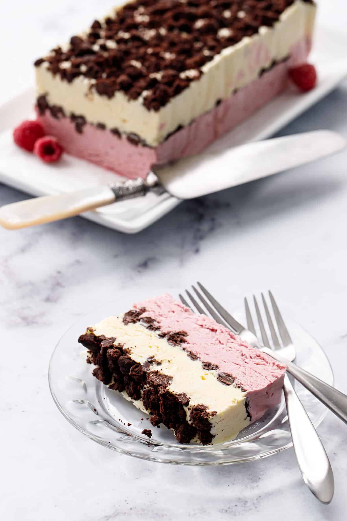 Slice of Raspberry & Passionfruit Semifreddo with Chocolate Crumbs on a glass plate, rest of the block of frozen semifreddo on a rectangle plate in the background.