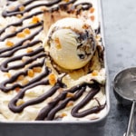 One round scoop sitting in a silver pan with Candied Orange Ice Cream with Chocolate Fudge Swirl, ice cream scoop on the side.