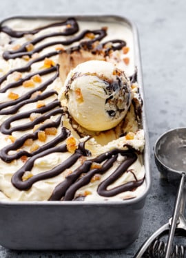 One round scoop sitting in a silver pan with Candied Orange Ice Cream with Chocolate Fudge Swirl, ice cream scoop on the side.